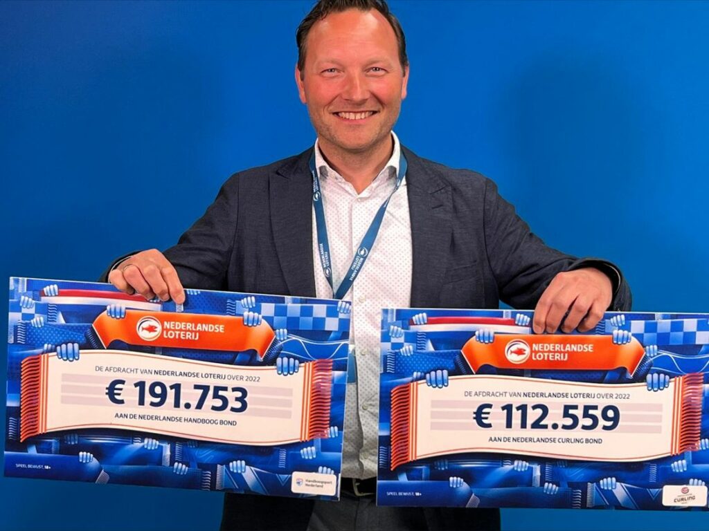 Arnoud Strijbis, directeur van de Nederlandse Curling Bond en de Nederlandse Handboogbond, houdt twee cheques vast voor een blauwe achtergrond. Voor elke bond één cheque.