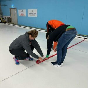 Drie vrijwilligers zijn op een witte ijsvloer bezig om rode tape uit te rollen langs een donkere, gespannen draad. Deze rode lijn komt onder het ijs en vormt dan de zogenaamde hogline in een curlingbaan.